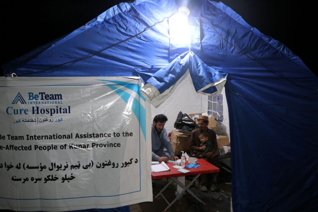 Be Team International Cure Hospital doctor listening to a father and young baby inside a temporary tent at a medical camp in Afghanistan at night