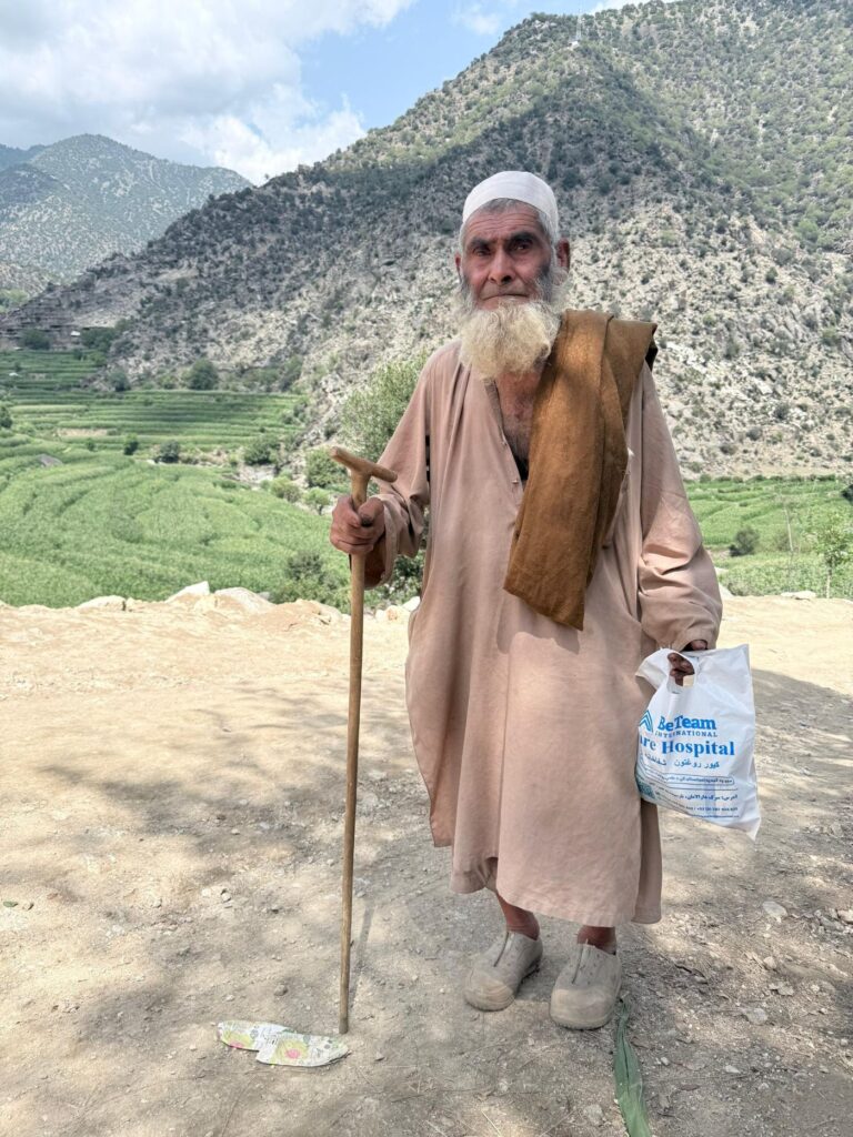 Elderly Afghan man holding medicine provided by Be Team International Cure Hospital team during a medical camp, with mountain range behind him