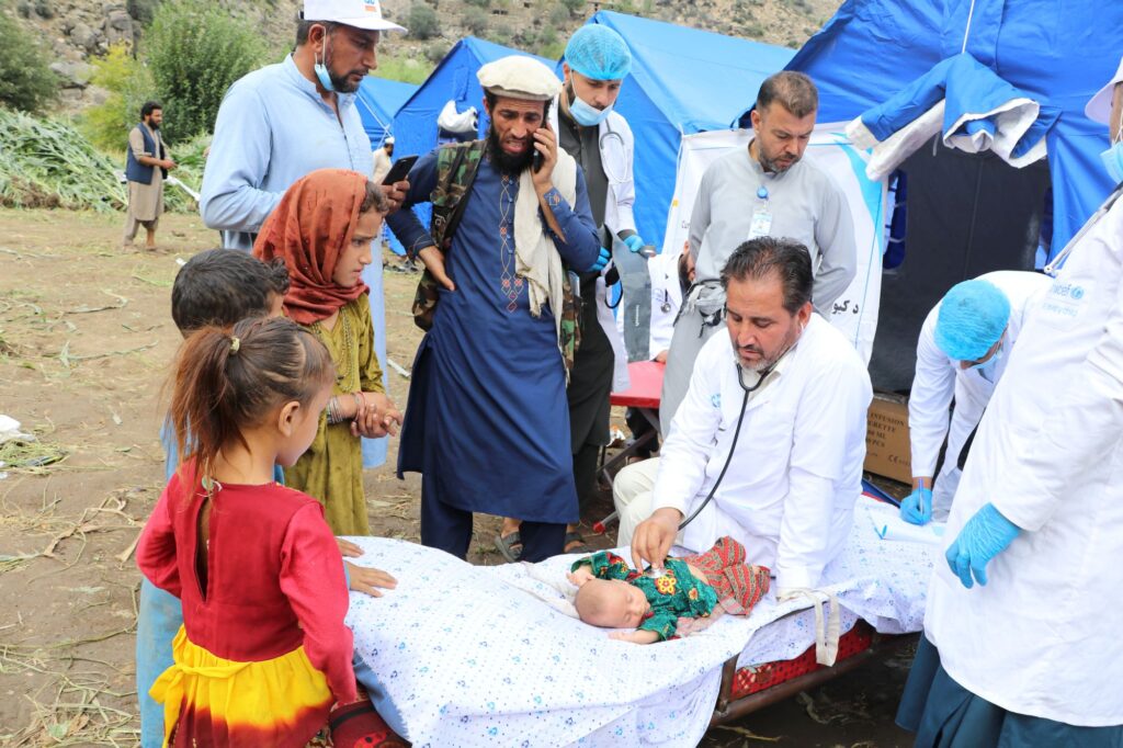 Be Team International Cure Hospital doctor examining a baby with a stethoscope at a crowded crisis response medical camp in Afghanistan