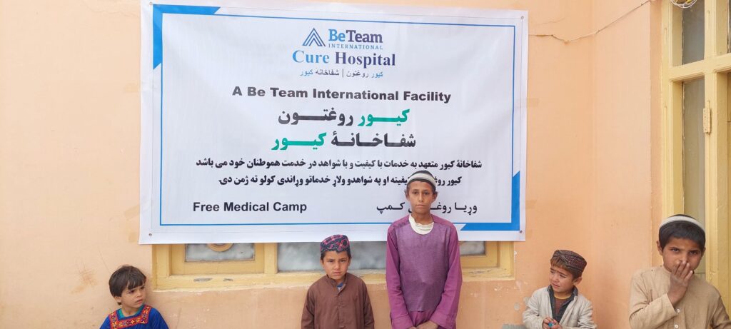 Four young Afghan boys standing by a poster advertising a free medical camp hosted by Be Team International's Cure Hospital in Laghman, Afghanistan