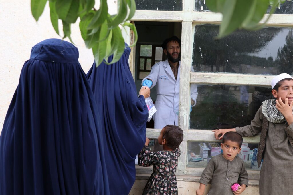 Be Team International Cure Hospital professional handing medicine through a window to an Afghan woman, as a young boy holds a flower nearby