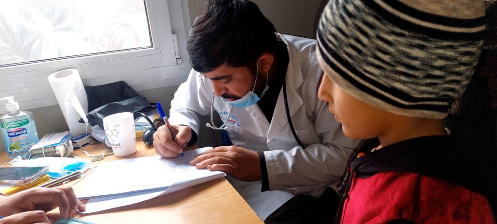 Be Team International Cure Hospital doctor conducting patient registration at a makeshift table during a medical camp in Tanic, Afghanistan, with a young boy watching nearby