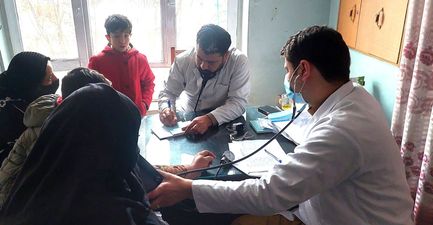 Doctors in a makeshift clinic examining a mother wearing a burqa in Afghanistan, her son watching nearby, at a Be Team International Cure Hospital outreach clinic